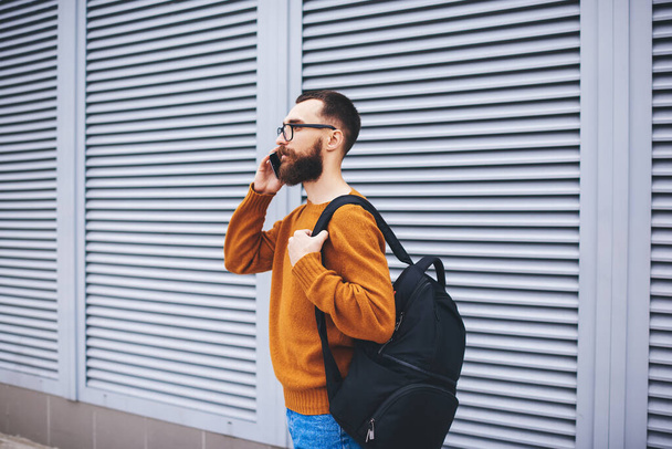 Side view of calm bearded man in stylish outfit with backpack looking away using smartphone for making call during break - Foto, Imagem