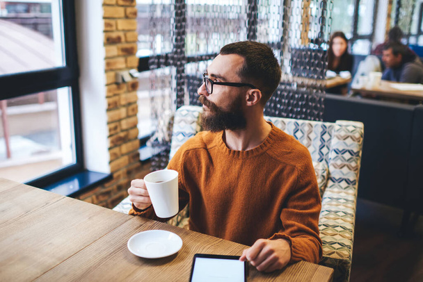 Pensive bearded man in casual outfit and spectacles looking away while enjoying cup of hot drink and browsing tablet in cozy cafe - Foto, afbeelding