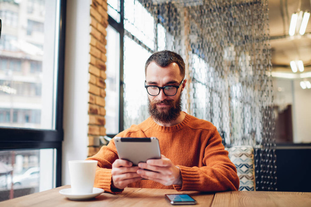 Positive unshaven male sitting at table with smartphone and cup of coffee while watching video on tablet chilling in light cafe - Foto, afbeelding
