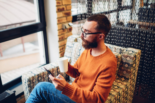 Widok z boku młodego przystojnego hipstera w modnym stroju i okularach o gorącym napoju i surfingu internet na telefon komórkowy siedzi w przytulnym fotelu w kawiarni - Zdjęcie, obraz