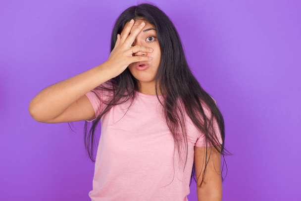 Ισπανίδα καστανή κοπέλα φορώντας ροζ t-shirt πάνω από μωβ φόντο peeking σε σοκ καλύπτει το πρόσωπο και τα μάτια με το χέρι κοιτάζοντας μέσα από τα δάχτυλα με αμηχανία έκφραση. - Φωτογραφία, εικόνα