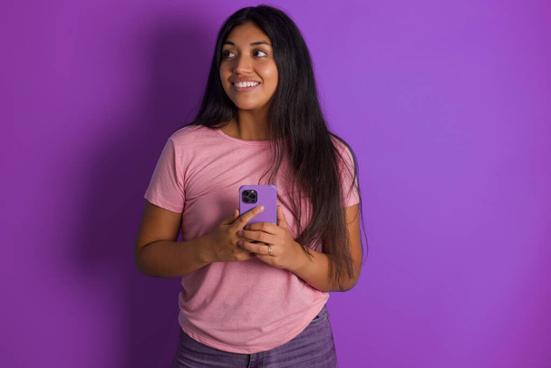 Hispanic brunette girl wearing pink t-shirt over purple background holding smartphone in hands - Photo, image