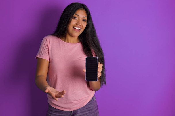 Spanisch brünettes Mädchen in rosa T-Shirt über lila Hintergrund präsentiert Smartphone. Werbekonzept. - Foto, Bild