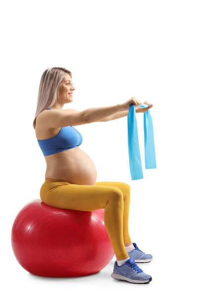 Full length profile shot of a pregnant woman sitting on a fitness ball and exercising with a resistance band isolated on white background - Foto, Imagem
