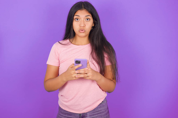 Shocked hispanic brunette girl wearing pink t-shirt over purple background opens mouth, holds phone, reading advert with unbelievable big shopping prices - Photo, Image