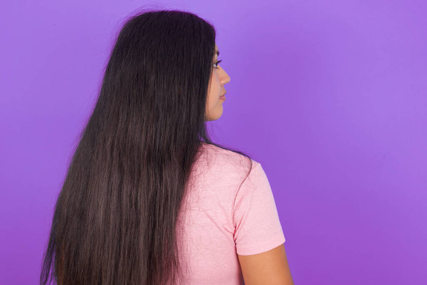 Takapuolella näkymä latino brunette tyttö yllään vaaleanpunainen t-paita violetti tausta. Studion kuvaus. - Valokuva, kuva