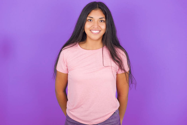 紫色の背景にピンクのTシャツを着たヒスパニックなブルネットの少女は,素敵な笑顔を喜んで表現しています. ポジティブな感情コンセプト - 写真・画像