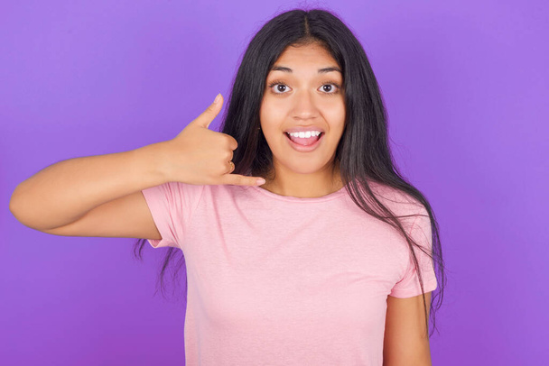 Молода іспаномовна дівчина в рожевій футболці над фіолетовим фоном робить телефонний жест, каже, передзвони мені знову, має радісний вираз
.  - Фото, зображення