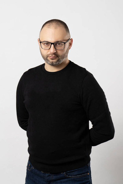 A young man with short hair and black-rimmed glasses holds his hands behind his back looking at the camera and poses on a white background - Foto, Bild