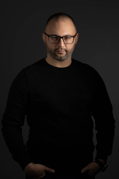 A young man in black-rimmed glasses and a black sweater keeps his hands in his pockets posing for the camera - Foto, immagini