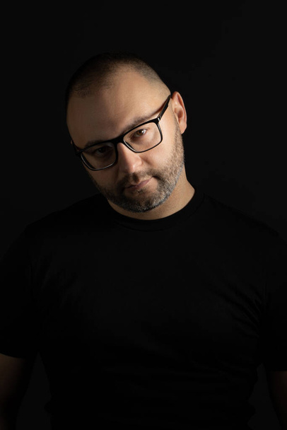 A young man in a black T-shirt with a short haircut and black-rimmed glasses poses against a dark background carefully and aggressively looking at the camera - Фото, изображение