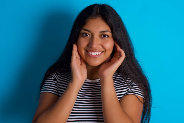 Happy Young hispánská dívka nosí pruhované tričko přes modré pozadí dotýká obě tváře jemně, má něžný úsměv, ukazuje bílé zuby. - Fotografie, Obrázek
