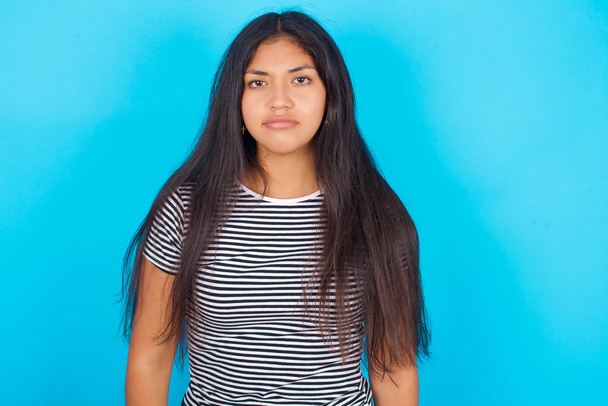 Nespokojenost Mladá hispánská dívka v pruhovaném tričku přes modré pozadí zamračí tvář cítí nešťastný má nějaké problémy. Koncept negativních emocí a pocitů - Fotografie, Obrázek