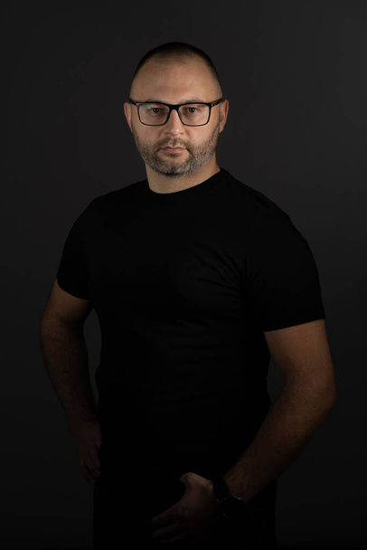 Un giovane uomo adulto con occhiali neri e una maglietta nera posa su uno sfondo scuro, guardando attentamente la fotocamera - Foto, immagini