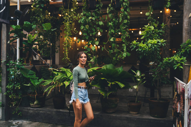 Side view slim pretty female in denim shorts looking away contentedly and standing outside rural house with open terrace decorated with green potted plants - Фото, зображення