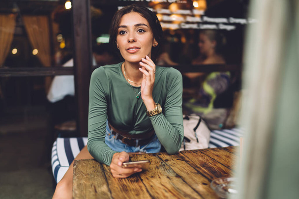 Young attractive female in casual clothes sitting at wooden table and holding mobile phone while spending time in cafeteria and looking away - 写真・画像