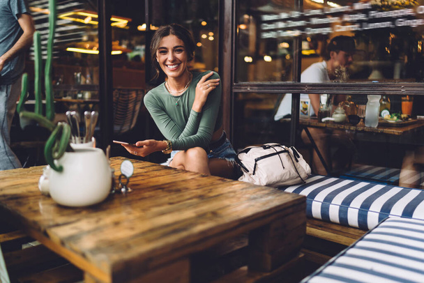 Cheerful female in casual clothes sitting at table and smiling while text messaging on mobile phone and looking at camera - Foto, afbeelding