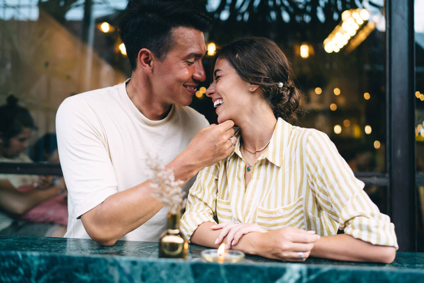 Happy young ethnic guy smiling and touching chin of laughing girlfriend while spending time together during romantic date in cozy cafe - Фото, изображение