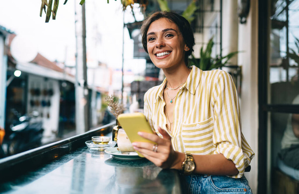 Happy young ethnic lady in stylish outfit leaning on counter and drinking cold cappuccino while sitting on cafe terrace with mobile phone - Zdjęcie, obraz