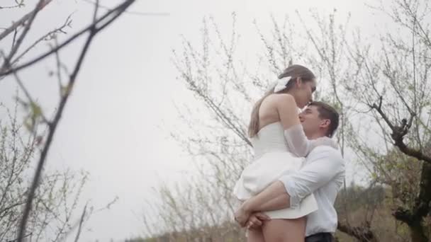 Beautiful couple embraces in spring garden. Action. Couple of newlyweds enjoy first leaves and flowers in garden in spring. Couple embraces in blooming garden in early spring - Filmati, video
