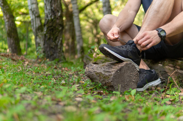 Man tying his shoelaces in the park outdoors, around the forest, oak trees green grass young enduring athletic athlete. run sport nature, lifestyle outdoor legs marathon, trees man. Summer body cross - Фото, зображення