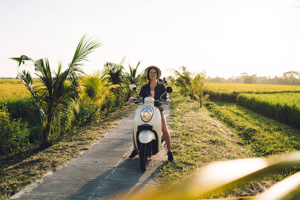 Portrait of attractive Caucaisan tourist posing at old fashioned scooter enjoying sunny weather at nature environment with path for driving, beautiful hipster girl on moped looking at camera - Photo, image
