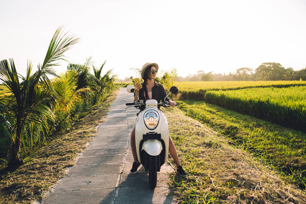 Attractive female travel blogger testing vintage motor transport during summer vacations holding cellphone device for networking, beautiful hipster girl with smartphone gadget resting on scooter - Photo, image
