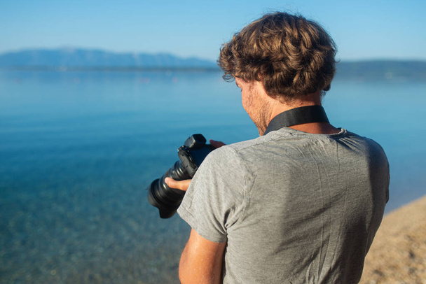 Widok z tyłu samca fotografa sprawdzającego i ustawiającego wyświetlacz aparatu stojącego nad morzem. - Zdjęcie, obraz
