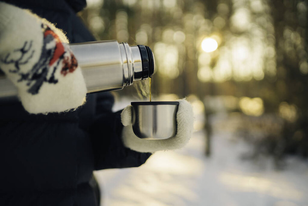 Woman traveler pours hot tea from a thermos into a mug, walking in snowy frozen winter forest. Adventure, travel, tourism and camping concept - Photo, image
