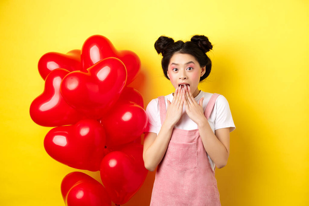 Έκπληκτο κορεάτικο κορίτσι λαχανιάζει και λέει wow, κοιτάζοντας ειδική προσφορά promo την ημέρα του Αγίου Βαλεντίνου, στέκεται κοντά μεγάλα μπαλόνια κόκκινες καρδιές και κίτρινο φόντο - Φωτογραφία, εικόνα
