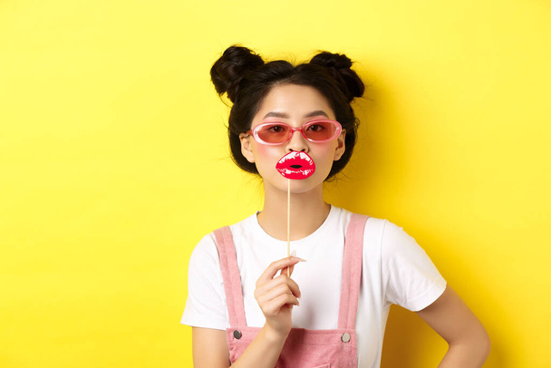 Concepto de verano y moda. Elegante asiático glam chica en gafas de sol, celebración de partido labios máscara, de pie sobre fondo amarillo - Foto, imagen