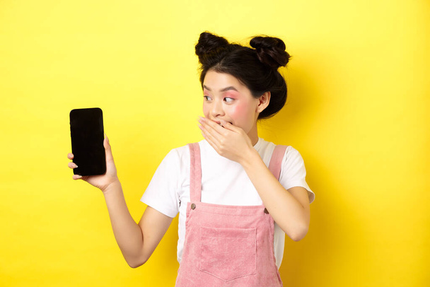 İnternetten alışveriş konsepti. Aptal Japon kız güzellik makyajlı, ağzı elle kaplı boş akıllı telefon ekranı göstererek gülüyor, telefonda komik bir şey gösteriyor, sarı arka planda. - Fotoğraf, Görsel