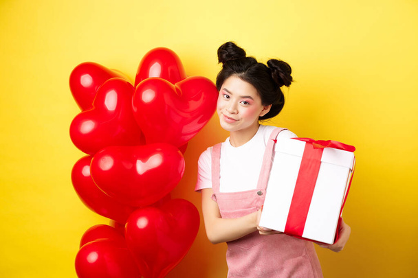 Ευτυχισμένη ημέρα του Αγίου Βαλεντίνου. Όμορφη και κομψή Ασιάτισσα γυναίκα θέτει κοντά μπαλόνια καρδιά με υπέροχο κουτί με δώρο από τον εραστή, στέκεται πάνω από κίτρινο φόντο - Φωτογραφία, εικόνα