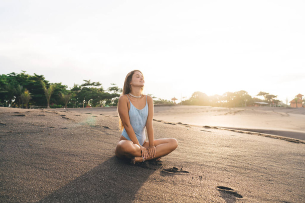 Full length relaxed young female wearing white swimsuit sitting with eyes closed in lotus pose on vast sandy shore against tropical greenery on early morning - Fotó, kép