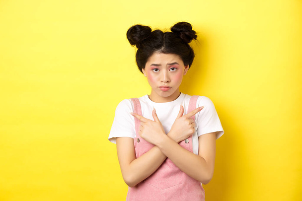 Indecisive timid asian girl pointing sideways, showing left and right ways and sulking upset, standing sad on yellow background - Photo, Image