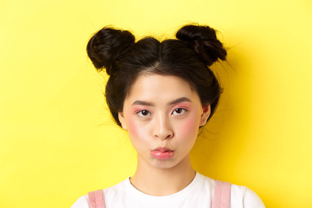 Close up portrait of moody asian girl pouting and looking upset at camera, standing wiht glamour makeup and hairbuns on yellow background - Photo, Image
