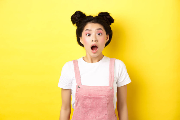 podekscytowany Azji nastolatek dziewczyna spadek szczęki, dysząc zdumiony z otwartymi ustami, patrząc na aparat pod wrażeniem, sprawdź lato promo oferta, stojąc na żółtym tle - Zdjęcie, obraz