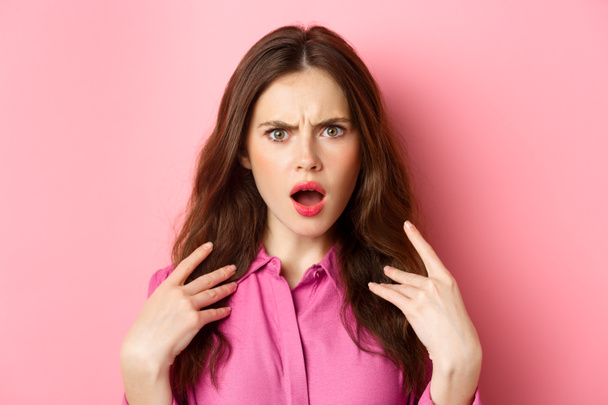 close-up van beledigd en geschokt jonge vrouw, fronsen en staren beledigd op camera, staande over roze achtergrond - Foto, afbeelding