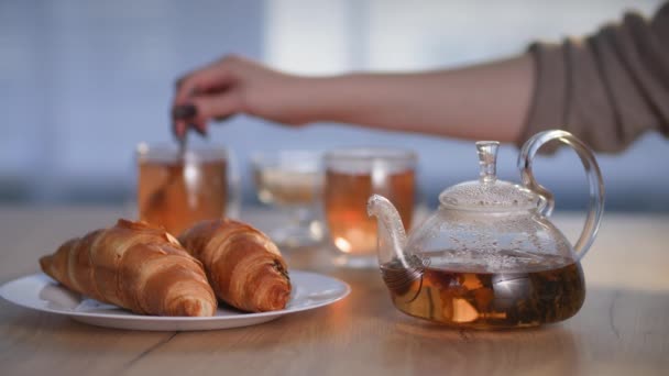 утренние ритуалы, травяной здоровый чай в чайнике с круассаном в тарелке, молодая женщина наливает сахар в чашку с горячим напитком сидя утром за столом во время завтрака - Кадры, видео