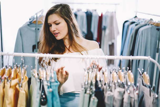 Focused woman in casual dress standing near clothes rails and choosing apparel while spending time in fashion store at daytime - 写真・画像
