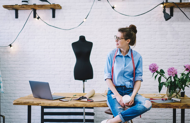Upset seamstress in glasses and casual clothes with takeaway hot beverage sitting on table with laptop and sketches in studio with mannequin - Foto, imagen
