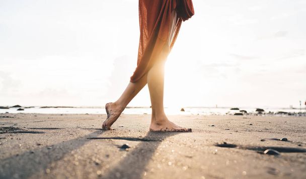Side view of crop barefoot faceless female in casual clothes walking on sandy beach near waving sea in sunny day on Bali - Фото, изображение