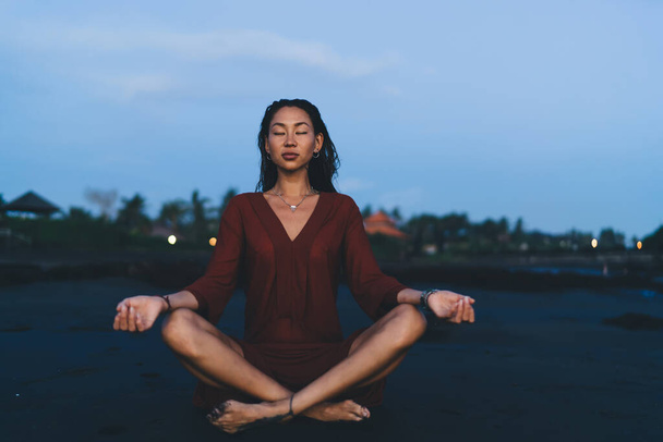 Slim Asian woman with closed eyes sitting on sandy beach and meditating while practicing lotus yoga pose during early morning - Фото, зображення