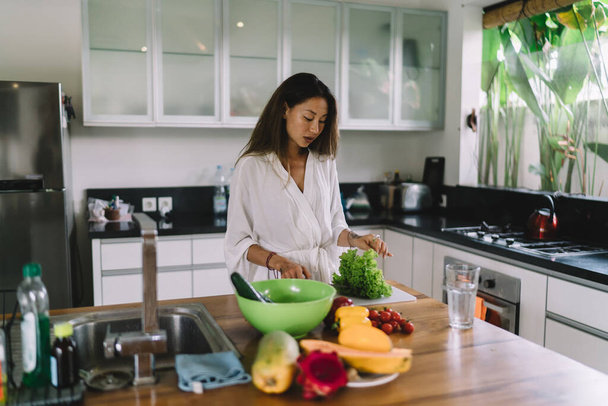 Concentrated young ethnic female with long dark hair in white robe cutting fresh lettuce while cooking vegetarian dish in stylish kitchen - Zdjęcie, obraz