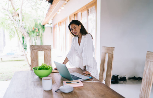 Smiling Asian freelancer with long brown hair in stylish clothes standing and leaning on table with laptop and bowl with fresh vegetables in light room in daytime - Foto, Imagem