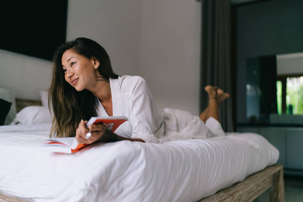 Charming Asian woman in white robe lying with book on comfortable bed and thoughtfully looking away in light bedroom in daytime - Fotoğraf, Görsel