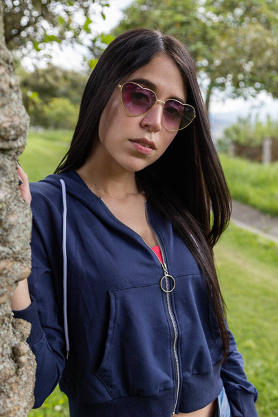 resting sitting on sunny day on the grass a young latin woman with straight black hair, wears jacket, jeans and sunglasses in fashion, lifestyle and beauty - Foto, immagini
