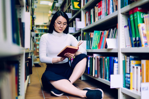 Moderní asijské studentky čtení knihy a kontrolu informací, zatímco sedí se zkříženýma nohama na podlaze vedle knihovny v univerzitní knihovně - Fotografie, Obrázek