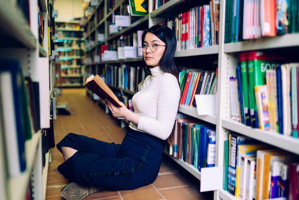 Dark haired Asian female with glasses in white turtleneck and blue trousers sitting cross-legged next to bookcase with opened book in hands and looking at camera - Foto, Bild
