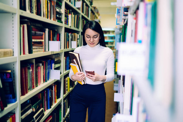 Молодая азиатская веселая студентка с красочными тетрадками в круглых очках ходит между книжными шкафами и печатает на смартфоне в библиотеке - Фото, изображение